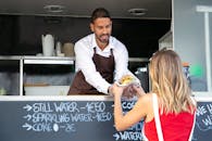 Positive ethnic cook in apron standing at counter in food truck and giving delicious hamburger to anonymous woman customer in daytime